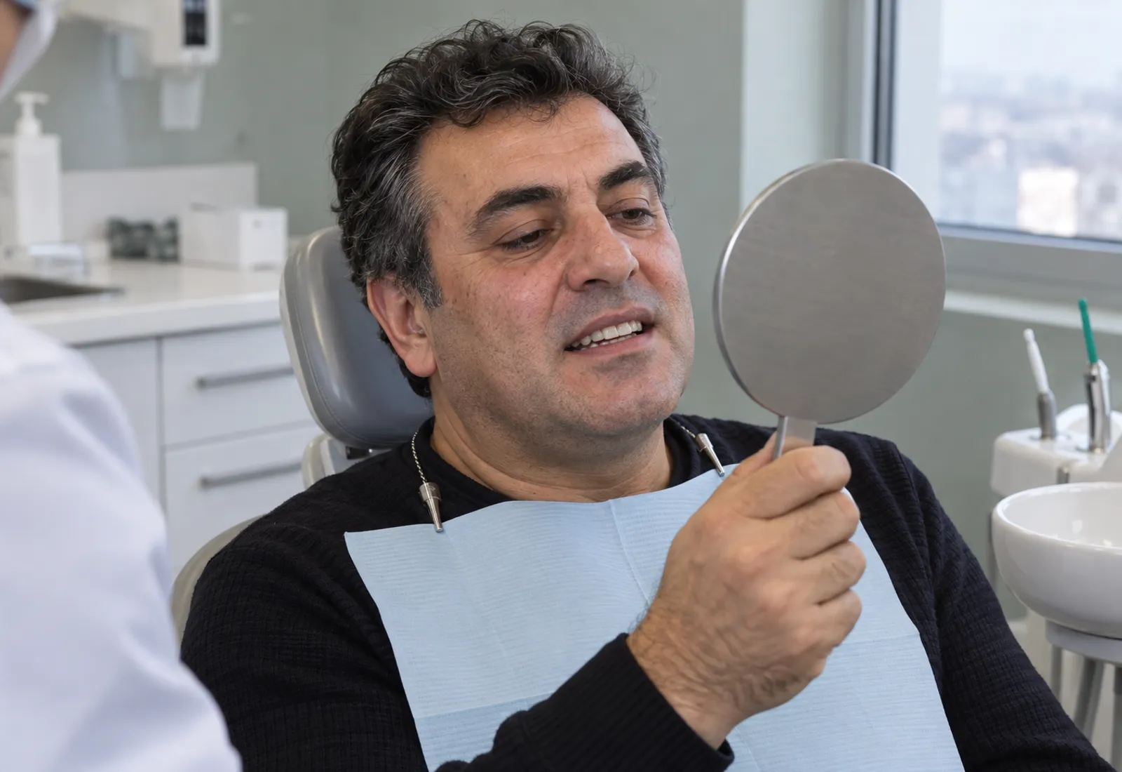 Diş koltuğunda oturan bir adam el aynasında yeni dişlerine bakıyor