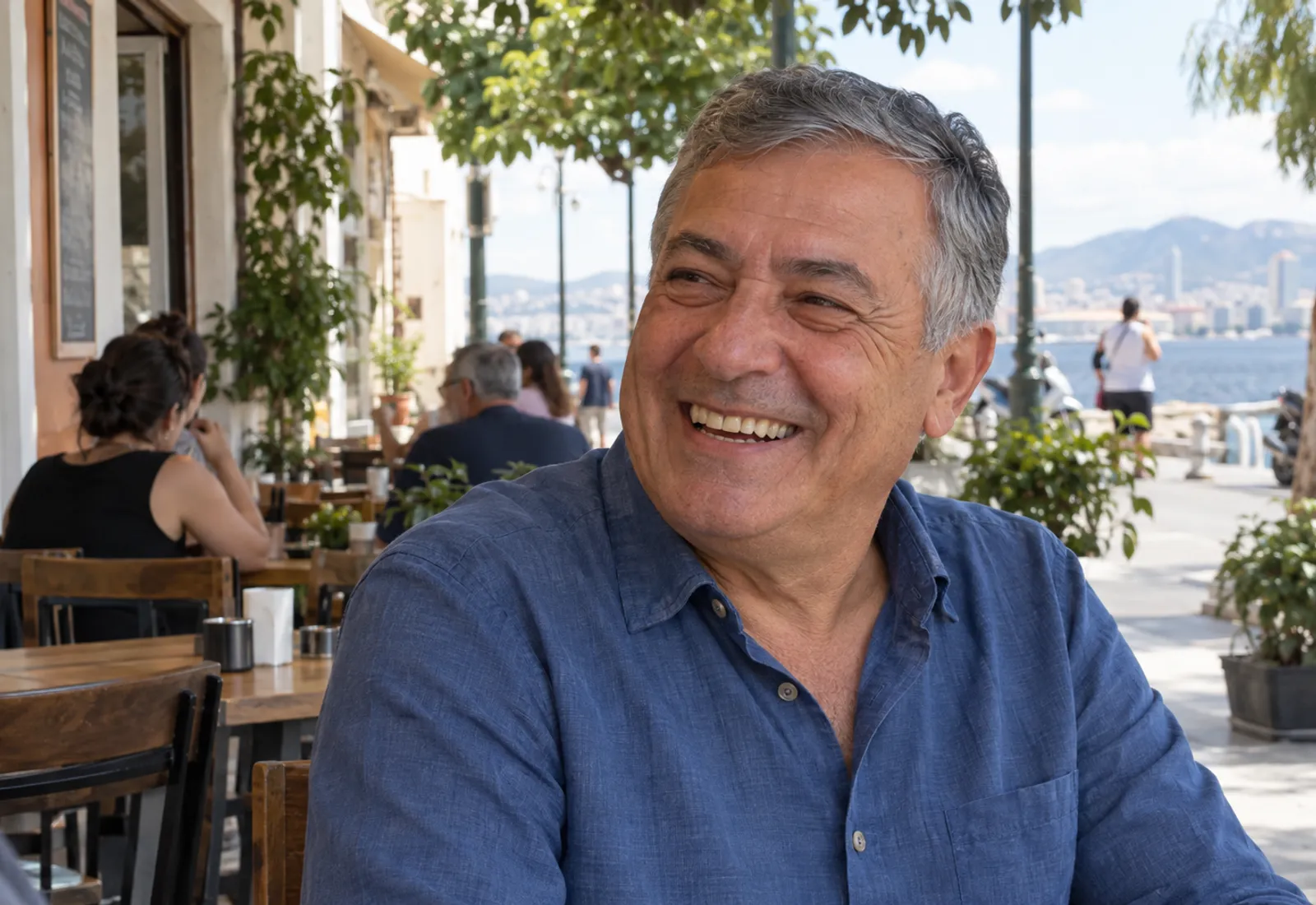 Altmışlı yaşlarında kır saçlı bir adam, deniz kenarındaki bir kafede sohbet ederken gülüyor